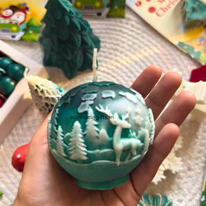 Teal decorative ball with deer and forest design held in a hand, with a blurred background of Christmas-themed items.