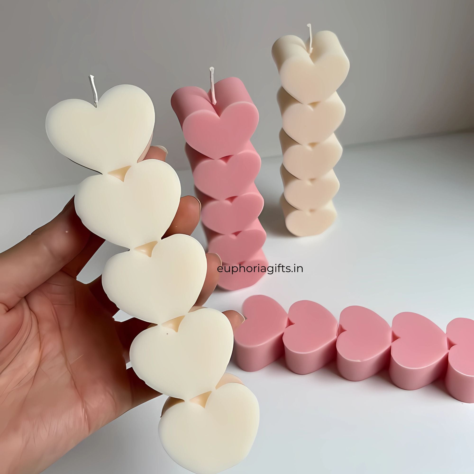 Heart-shaped candles in various colors held by a hand on a white surface with 'euphoriagifts.in'  Hand holding a white heart pillar candle with pink variants in the background, showcasing size and 3D sculptural quality branding.