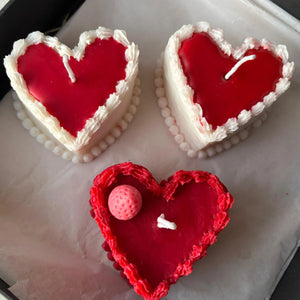 Hand holding heart-shaped cake candle made from natural soy wax, with red center and white frosting-like edges.