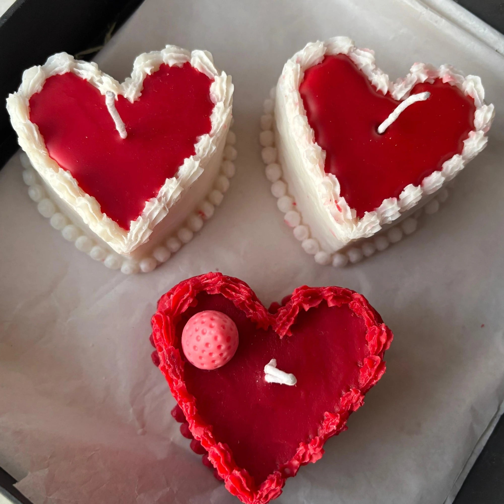 Hand holding heart-shaped cake candle made from natural soy wax, with red center and white frosting-like edges.