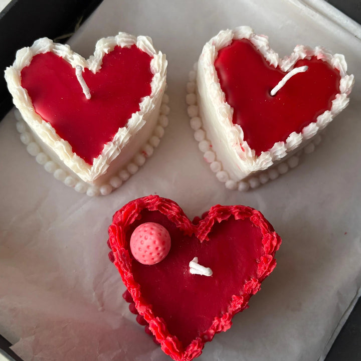 Hand holding heart-shaped cake candle made from natural soy wax, with red center and white frosting-like edges.