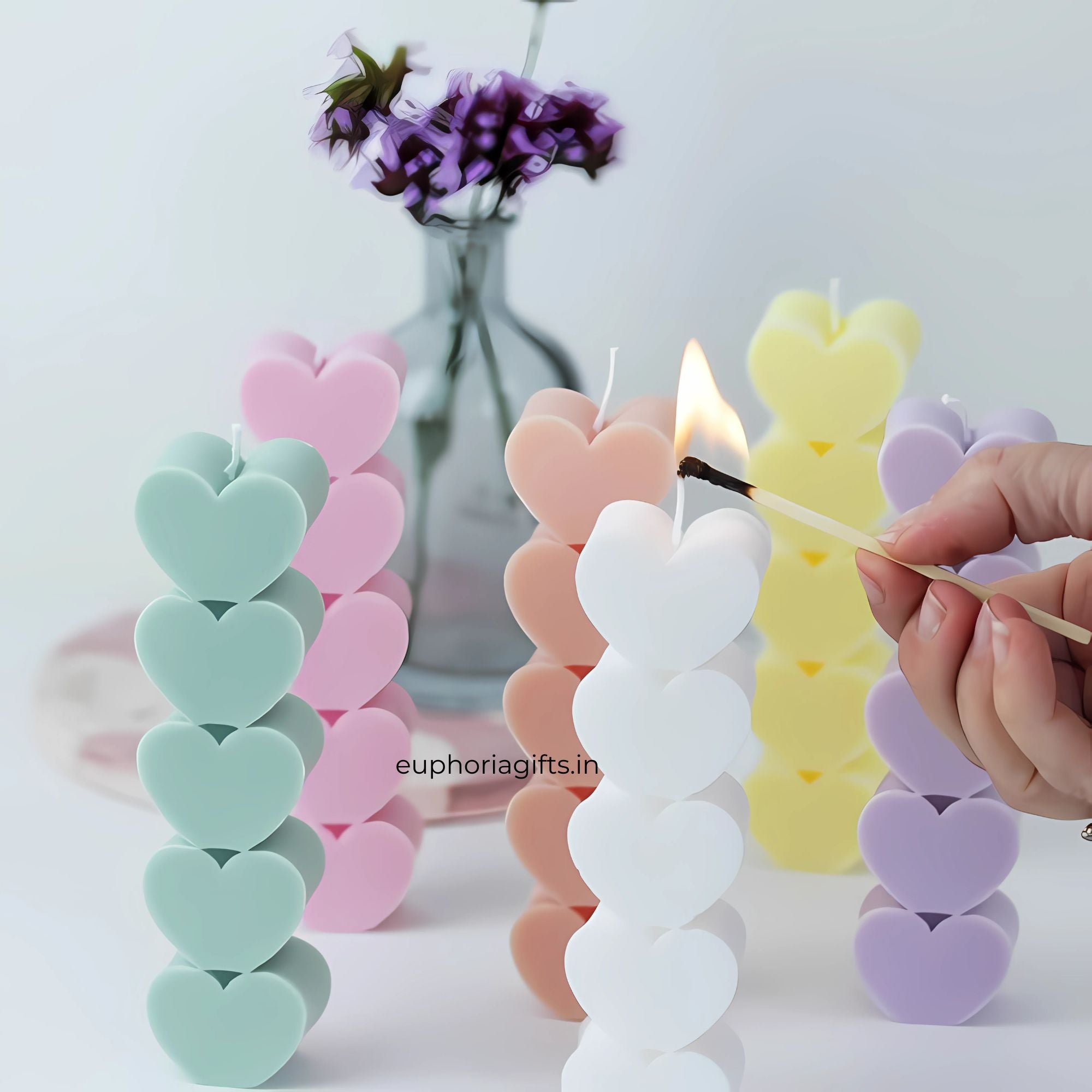 Heart-shaped candles being lit with a match, set against a light background. Assorted heart-shaped pillar candles in pastel colors being lit, set beside a vase with flowers for a cozy, aesthetic ambiance.